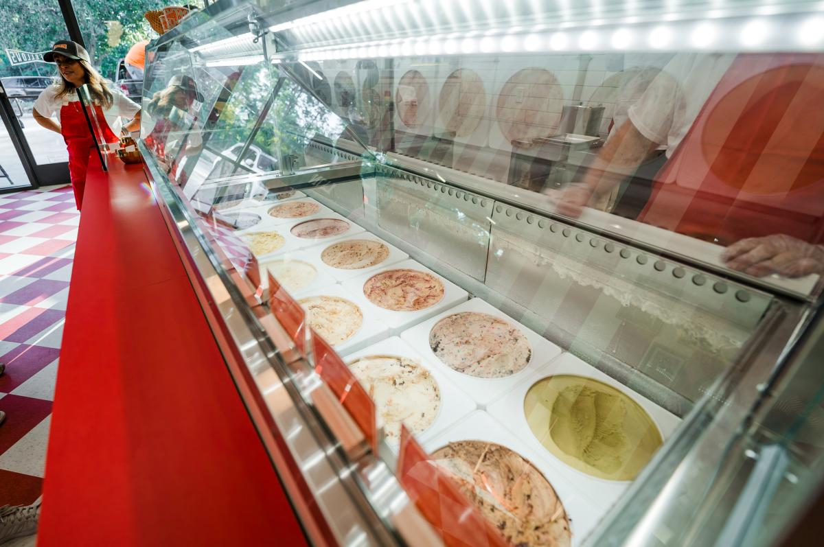 The camera looms over the ice cream counter to view all the flavors. A female employee stands at the end of the counter, speaking to customers standing out of the frame.