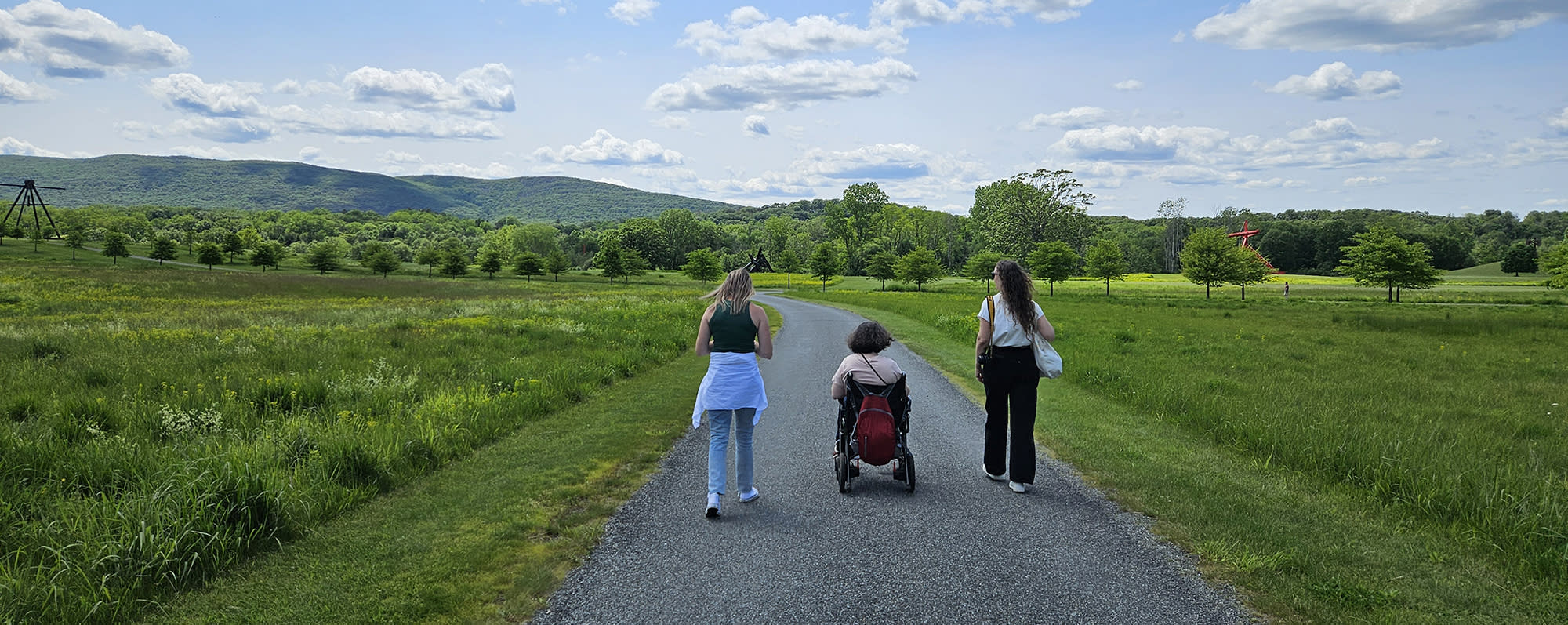 Three people including a person in a power wheelchair on a path surrounded by grassy fields at Storm King Art Center