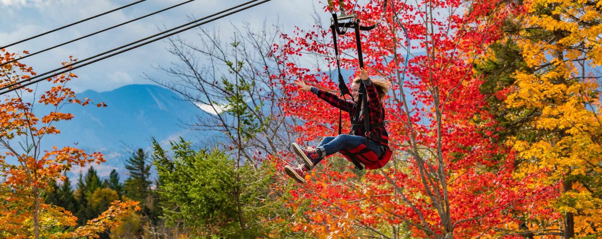 A woman with blonde hair and a red flannel shirt rides down a zip line at the Olympic Jumping Complex during peak fall foliage season