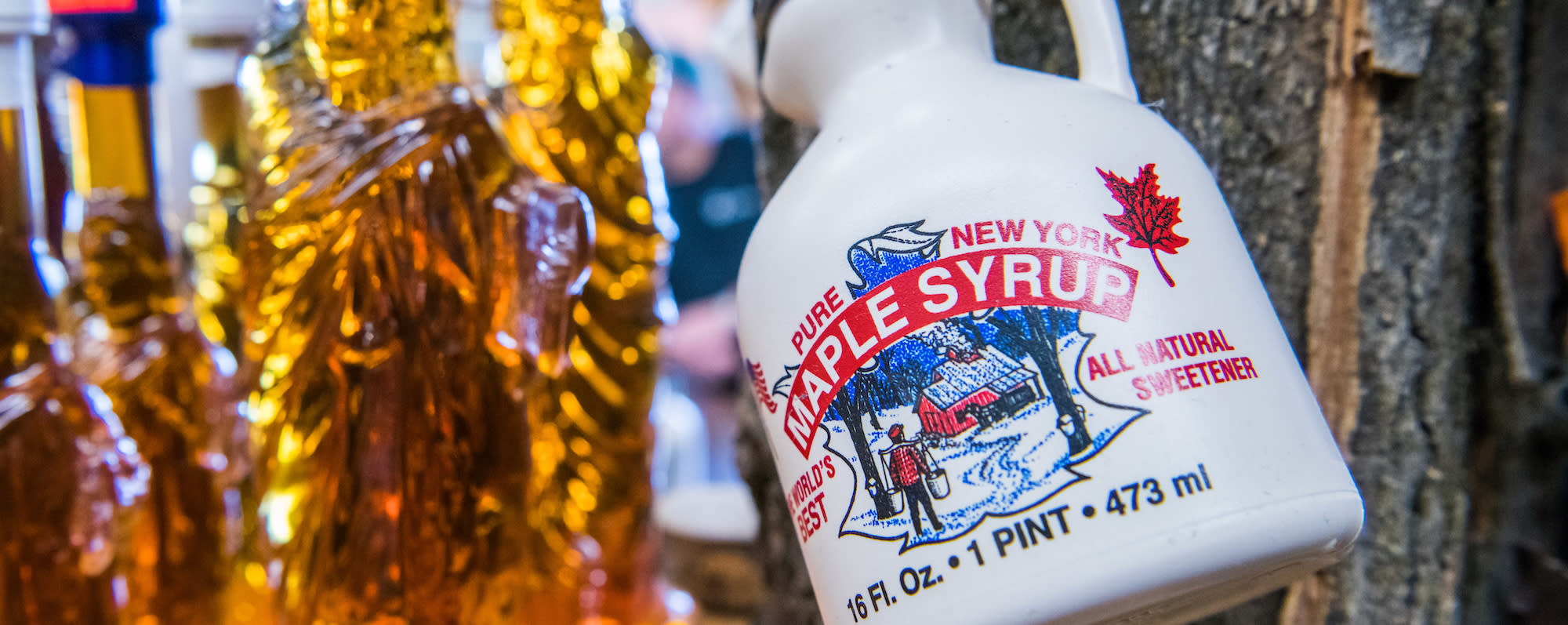 A bottle of New York Maple Syrup hangs on a tree with glass bottles of amber maple syrup sit nearby