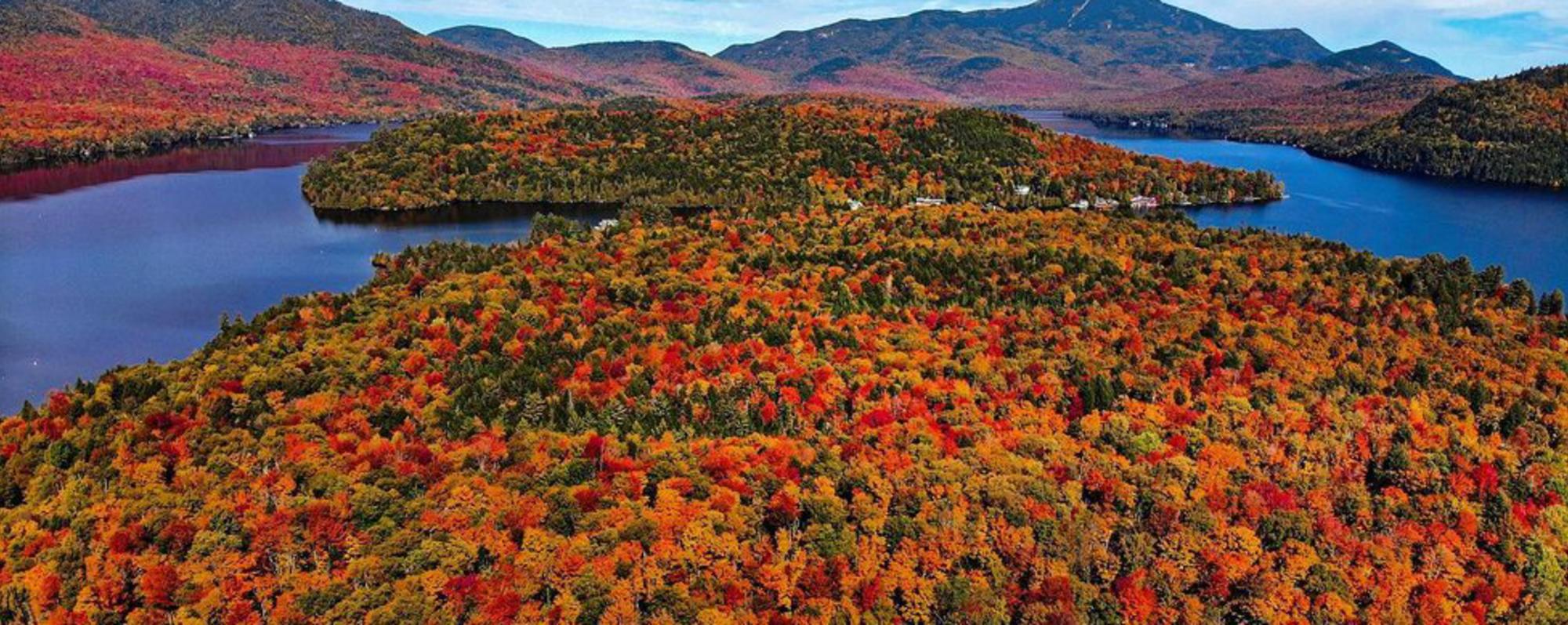 Varying colors of red, orange, yellow, and green trees surround blue lake waters with colorful mountains in the background