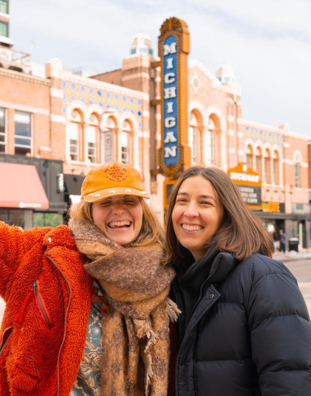 Two women smile as they pose in front of historic buildings, including the large "Michigan" sign that marks the Michigan Theater.