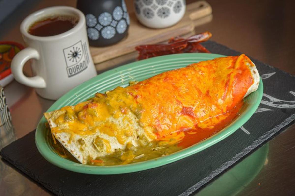 A photo of a breakfast burrito from Duran Central Pharmacy. The burrito sits on a turquoise plate. Half of it is smothered in green chile. The other half is smothered in red chile. A mug of coffee is in the background.