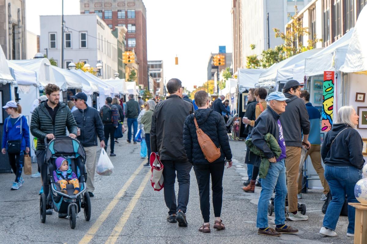 people walking both directions down a closed street lined with vendor tents full of artwork. People wearing coats and one man pushes a stroller with a baby resting.