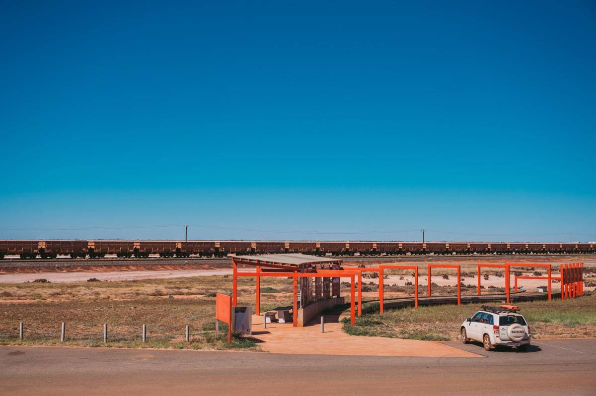 A car is parked at Redbank Bridge Lookout with a long iron ore train visible in the background. Alongside the carpark is a shaded lookout area with intrepretative signage.