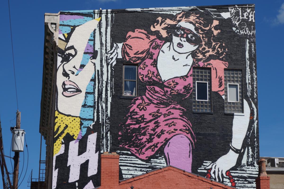 A street art mural by Faile in Covington, Ky. of a woman in a pink dress, wearing a black mask, climbing out of a building