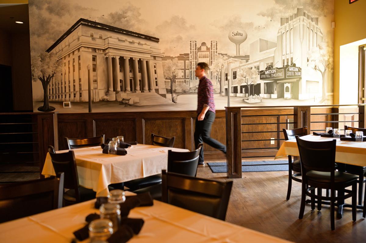 Inside of a restaurant with linen covered tables. A man walks in front of mural on a wall.