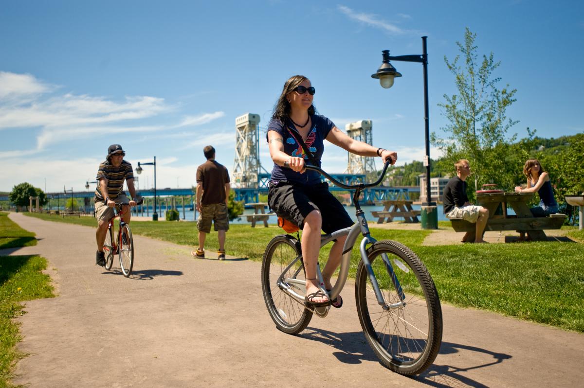 Bikers bike along Houghton waterfront with the bridge and people sitting at a picnic table in the background.