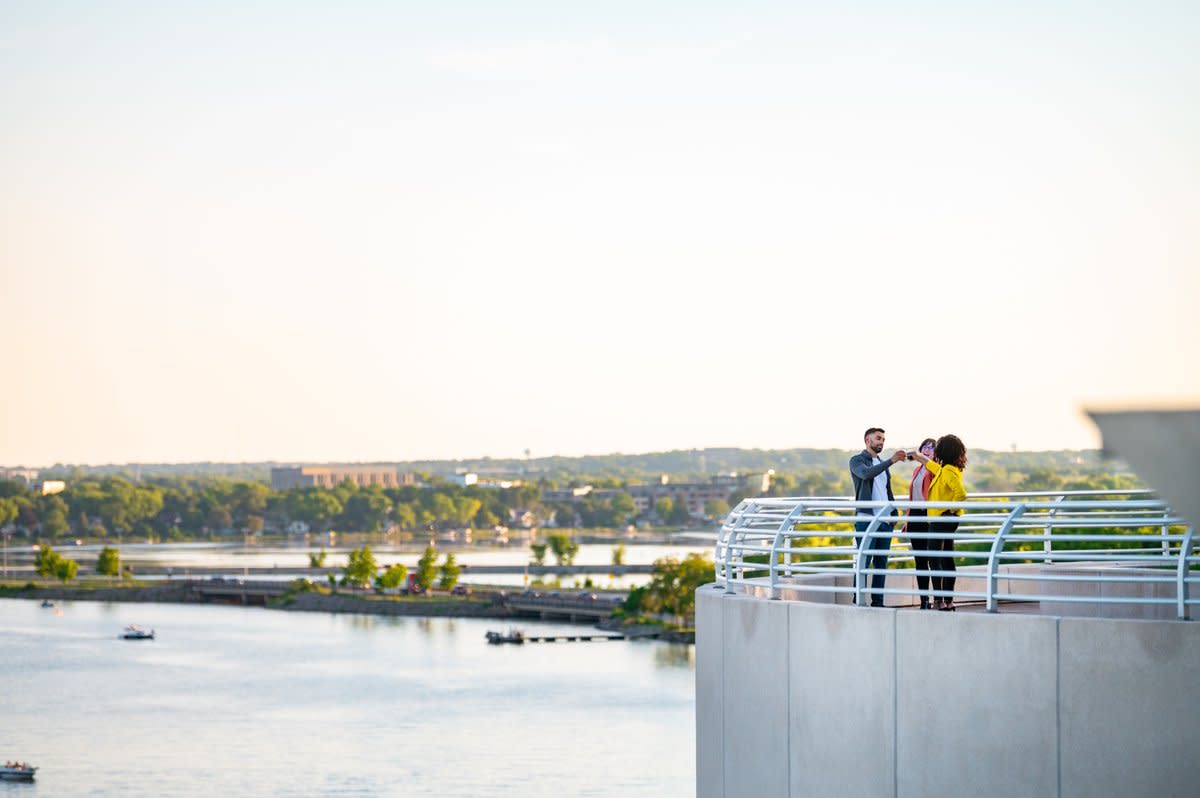 A group of three people chat and laugh on the rooftop of Monona Terrace with the lake and sunset in the background.