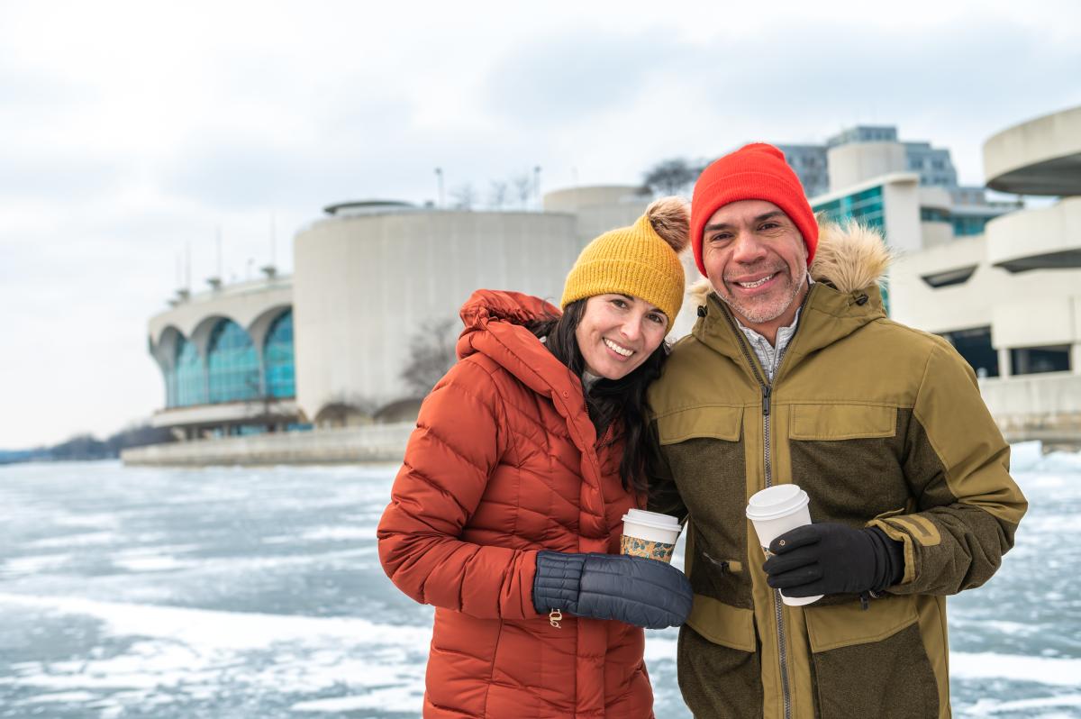 A white man and woman smile while holding coffee cups standing on a frozen Lake Monona in front of Monona Terrace.