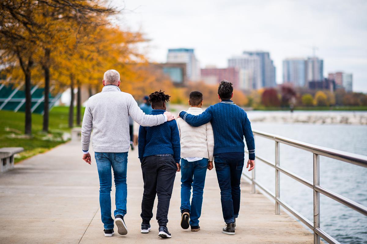 Two adults and two children walk arm-in-arm along Milwaukee’s lakefront path on a crisp fall day, with autumn trees on one side and the city skyline in the background.