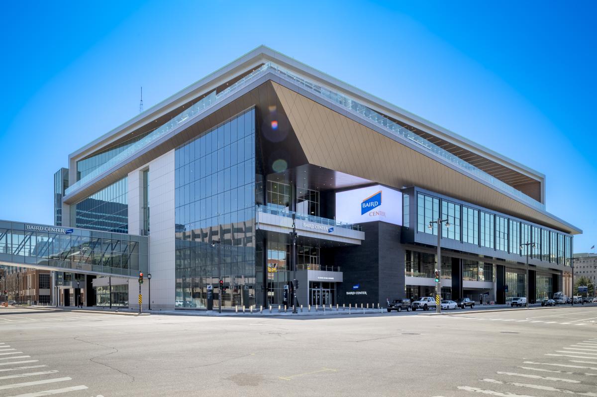 A daytime exterior view of the Baird Center in Milwaukee, showing its modern glass façade, angular roofline, and skywalk connection over the street, with cars and pedestrians nearby.