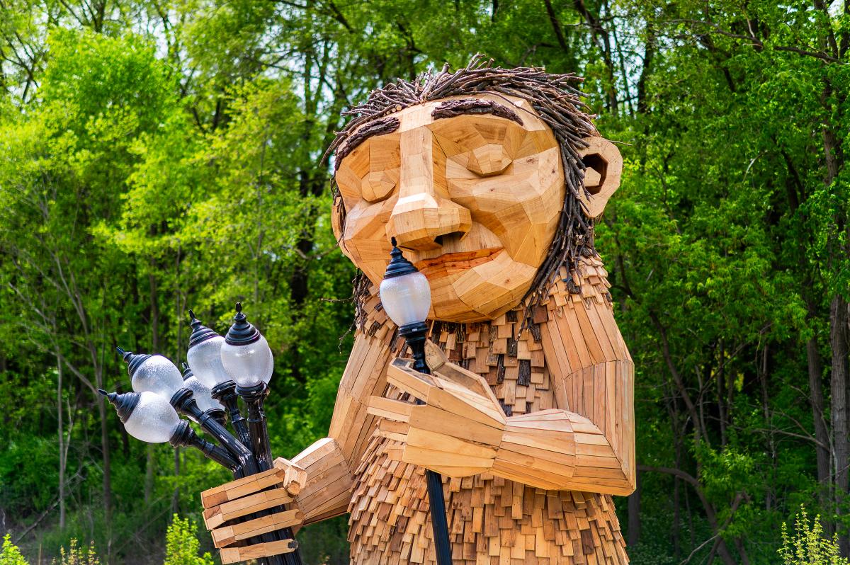 A large wooden sculpture of 'Mama Rosa,' a Thomas Dambo troll installation in Wauwatosa. The friendly giant figure is constructed from reclaimed wood pieces in natural tones, featuring a carved face with a gentle smile and twig-like hair made from bundled branches. The troll holds a bouquet of old-fashioned street lamps in one hand while lifting one lamp to her nose. The sculpture is set against a lush green forest backdrop with dense tree canopy, creating a whimsical scene that blends art with nature.