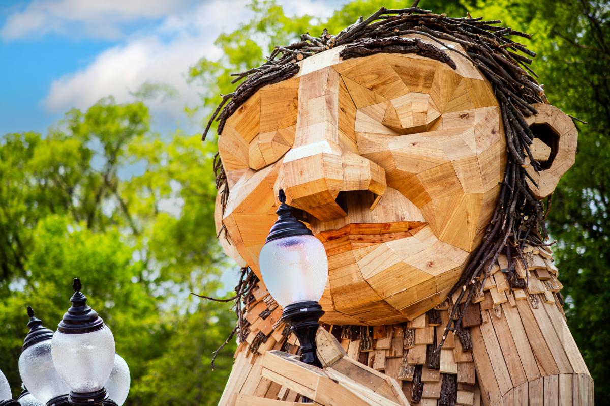 A large wooden sculpture of a humanoid figure, made from planks and branches, holds a lamp post close to its mouth as if about to take a bite. The figure has twig hair, a geometric facial structure, and is surrounded by green trees and a partly cloudy sky.