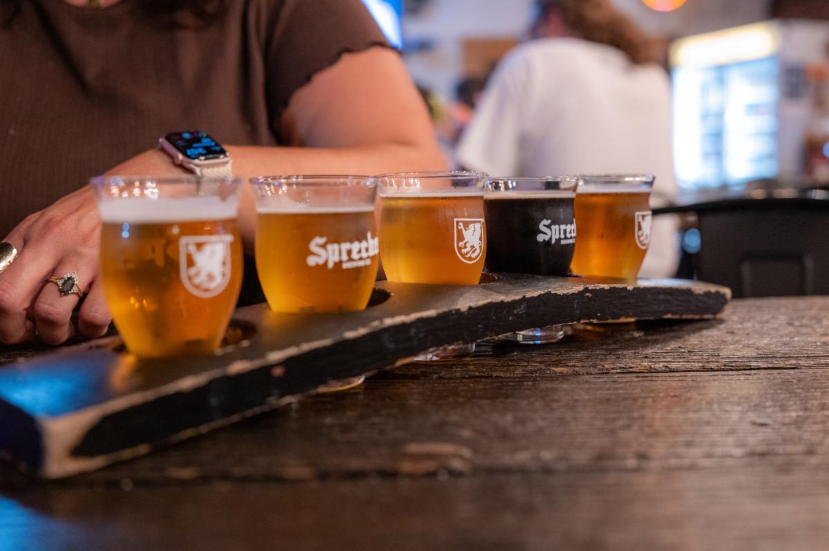A flight of five Sprecher beers, ranging from golden ales to dark brews, sits on a wooden tasting paddle at a brewery.