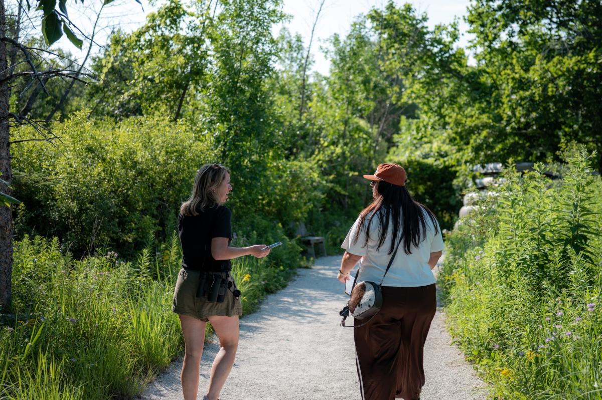 Two women walk along a gravel nature trail surrounded by lush greenery and wildflowers on a sunny day.