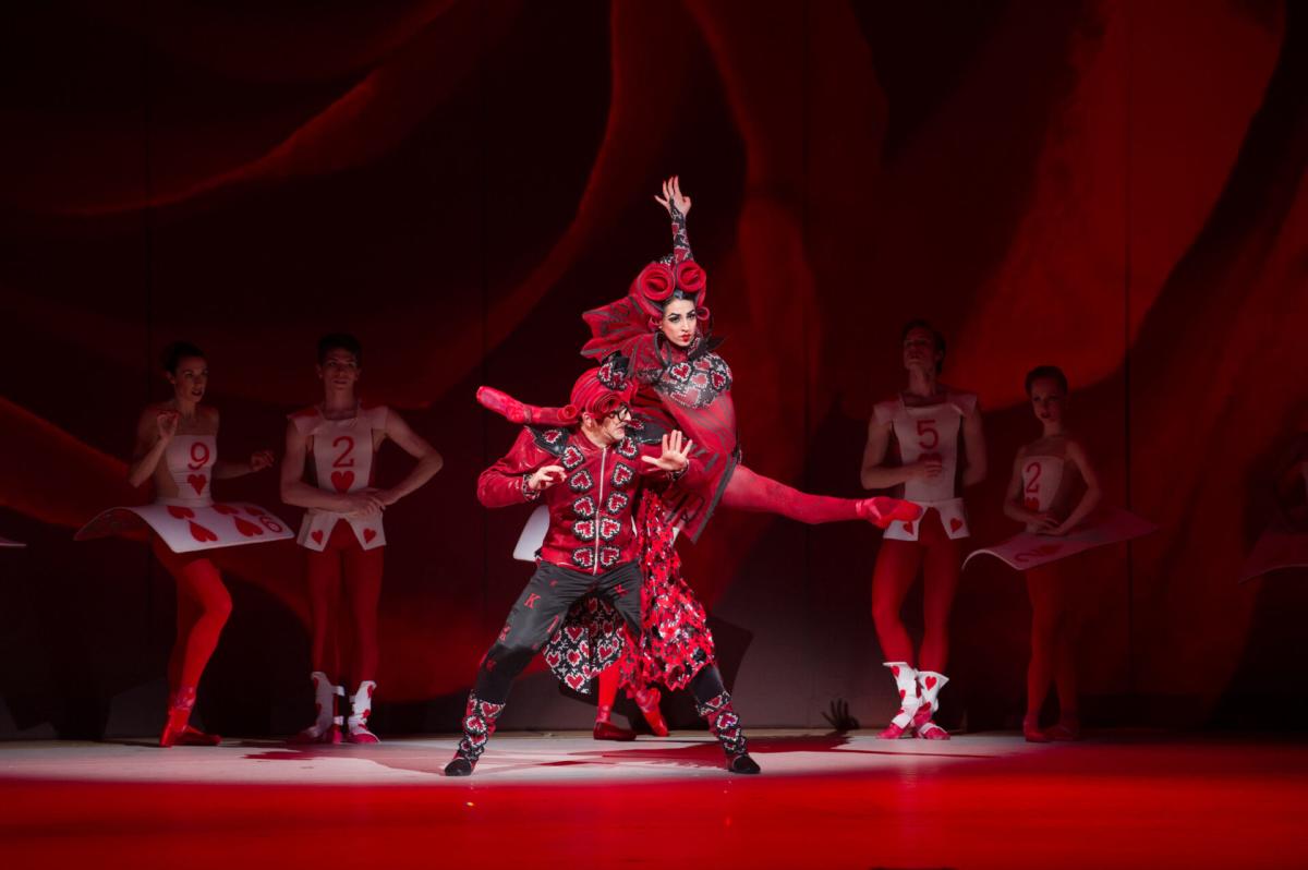 Dancers perform a scene from Alice in Wonderland, featuring the Queen and King of Hearts in red-and-black costumes at center stage. The Queen of Hearts is lifted into the air while the King of Hearts stands below. Other dancers dressed as playing cards stand behind them under red stage lighting.