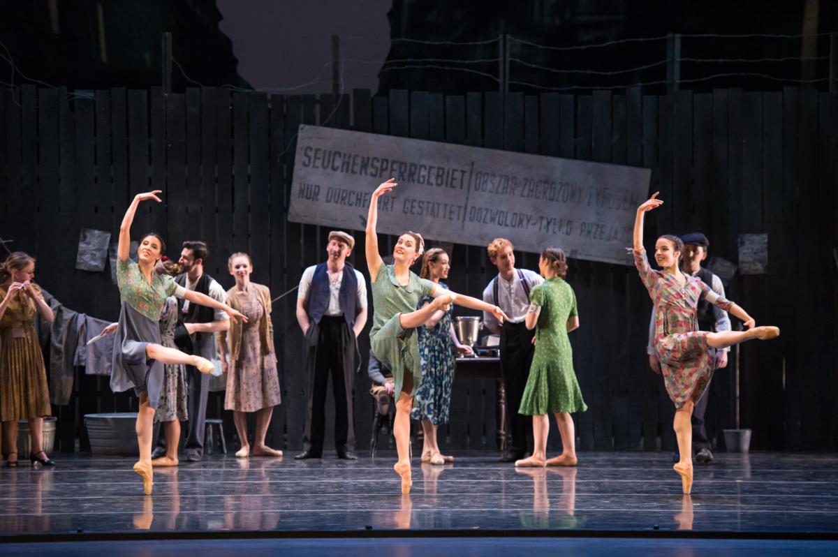 A group of ballet dancers perform onstage in vintage-style dresses, striking elegant poses with lifted arms and extended legs, while other performers in period clothing watch from behind.
