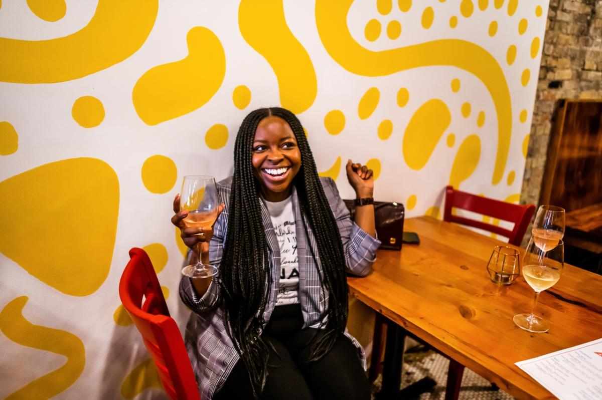 a woman sitting in front of a yellow and white patterned wall laughing and smiling while holding a glass of white wine