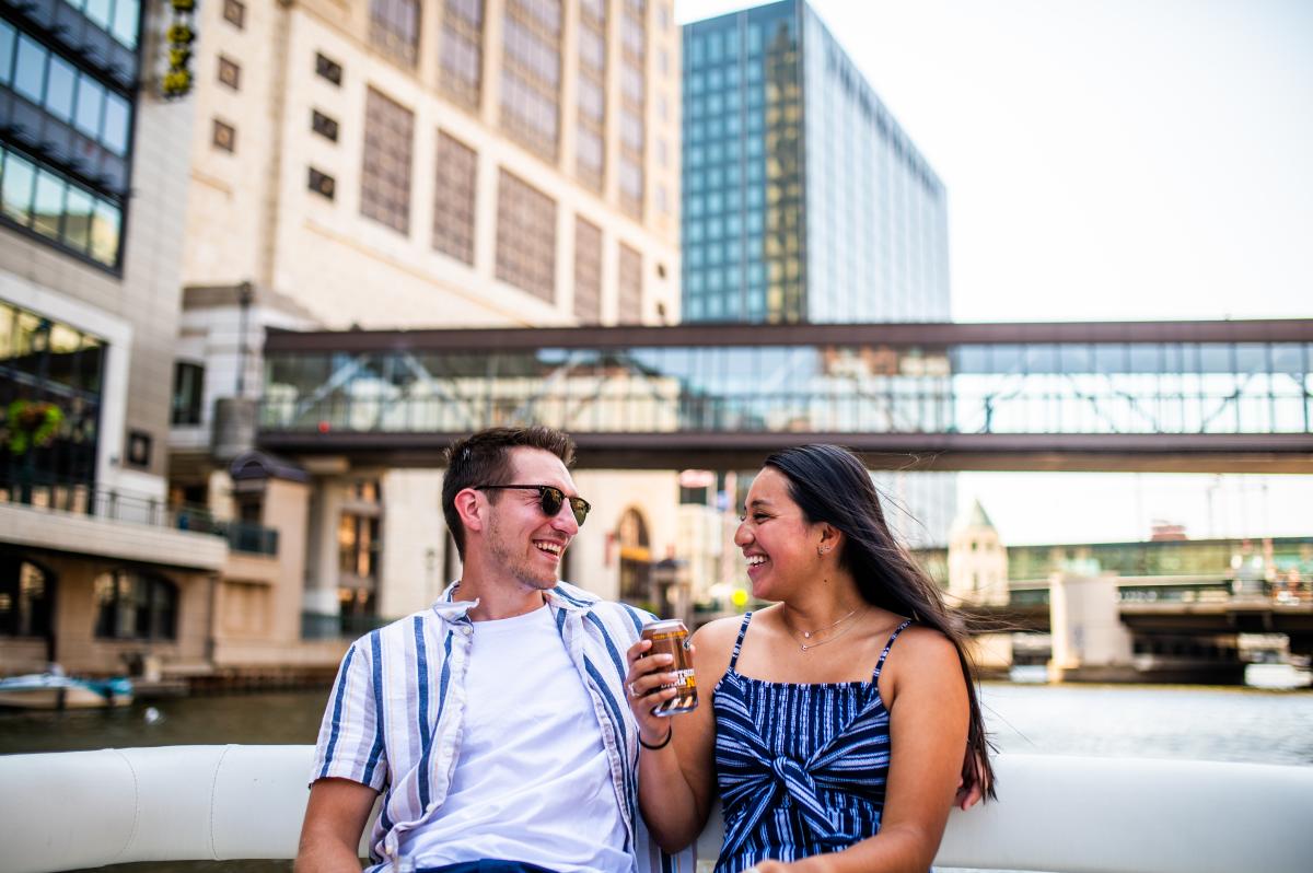 Couple laughing with each other on the back of a boat on the Milwaukee River with tall buildings in the background