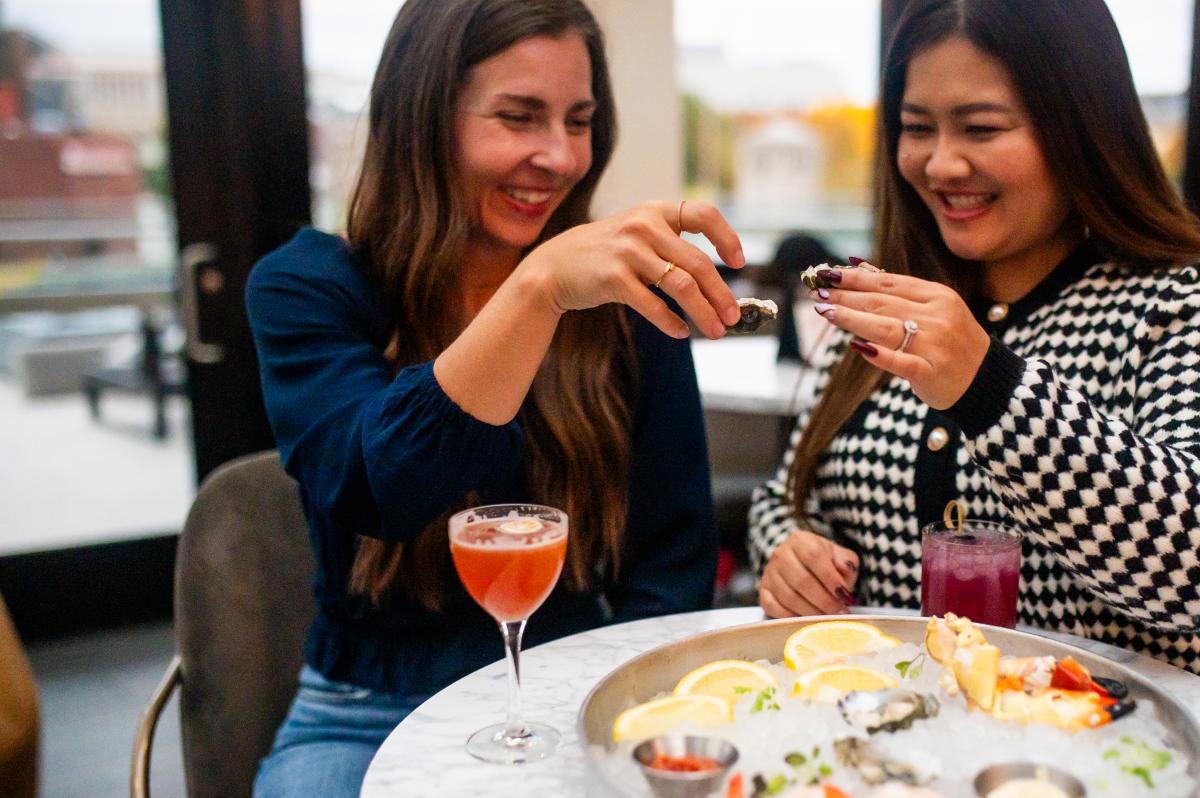 Two women smiling and toasting with oysters at a table featuring a seafood platter and cocktails, seated indoors with soft natural lighting.