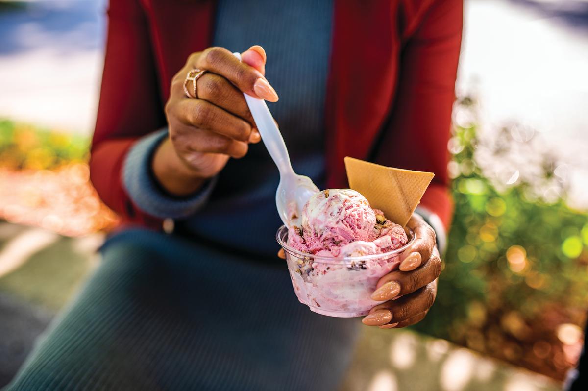 A person holds a cup of berry swirled frozen custard from Kopp's Frozen Custard, topped with a waffle cone wafer, while lifting a spoonful to eat. The close-up highlights the colorful dessert, with a softly blurred outdoor background.