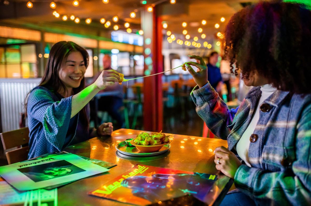 two women sitting at a table in a dim, string-light-lit bar pulling apart a cheese curd to stretch the cheese inside