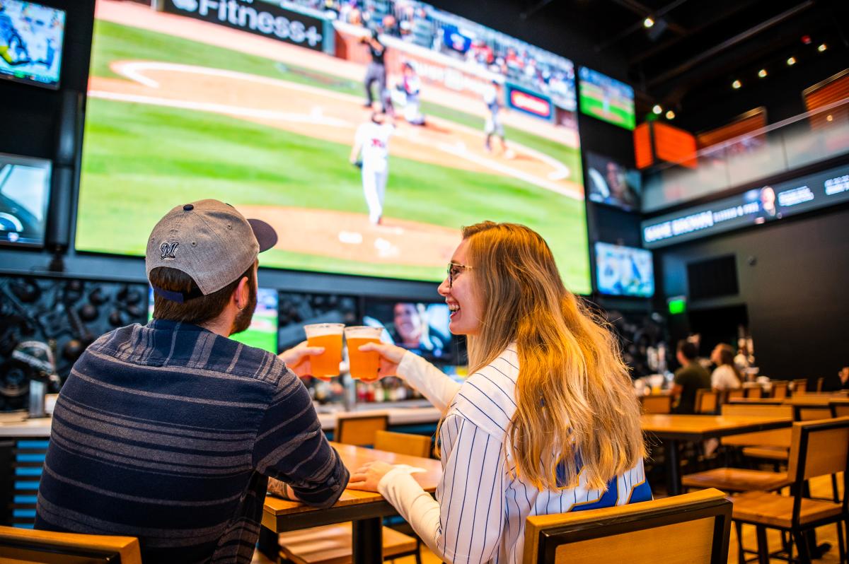 A man and woman clinking glasses of beer while watching baseball