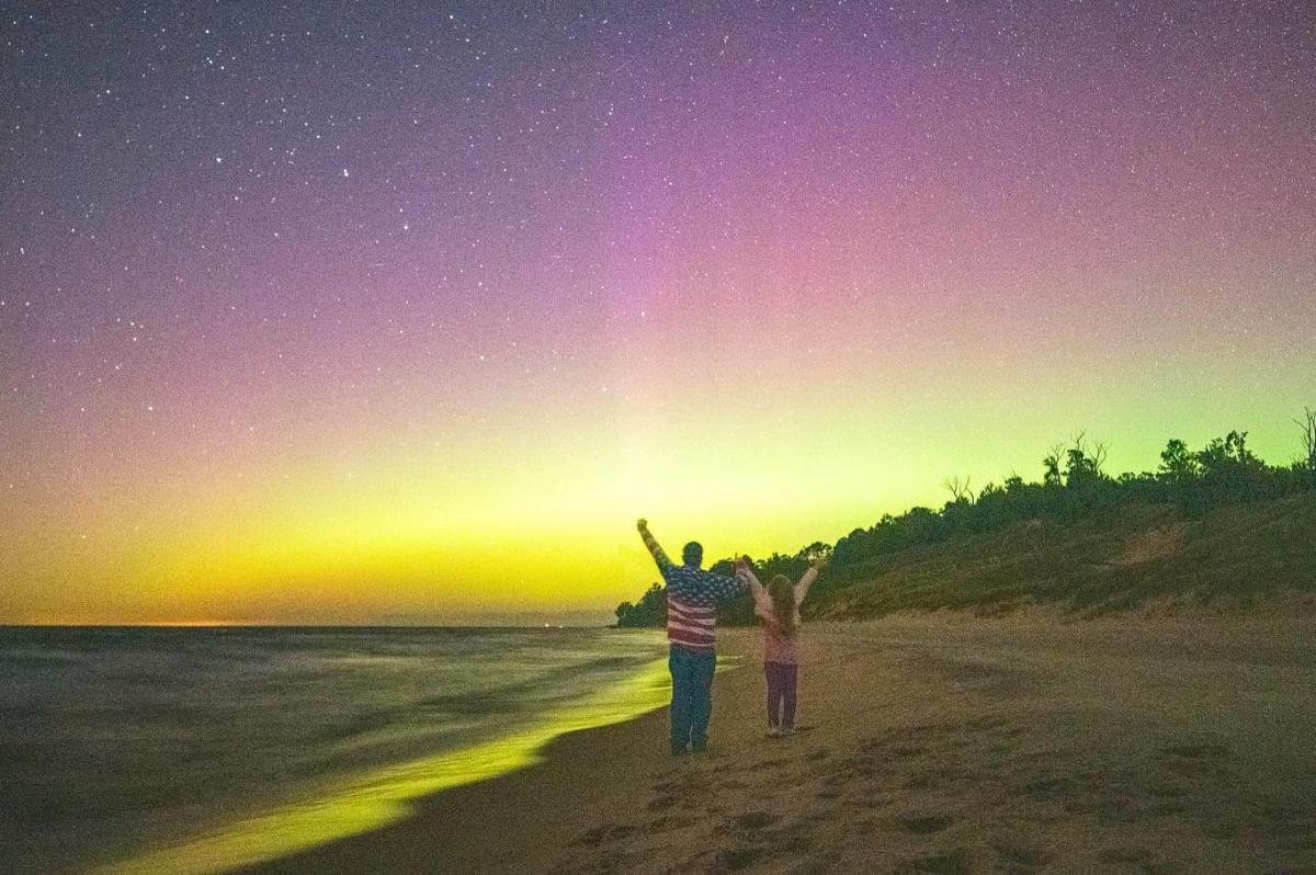 Two children standing on a Lake Michigan beach in Muskegon, Michigan at night, pointing at a sky illuminated by stars and the Northern Lights.