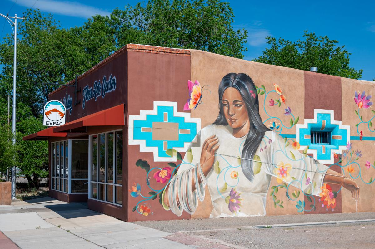 Mural of a person with long hair wearing traditional attire on the side of the Espanola Valley Fiber Arts Center in a sunny setting. The mural features bright floral designs and geometric patterns.