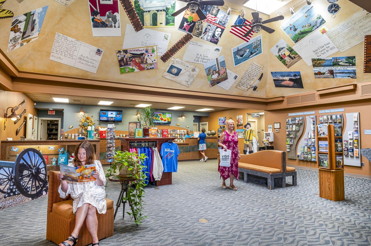A woman is sitting reading the visitor guide magazine while another woman shops in a vaulted room with an artistic ceiling covered in places around Newport News