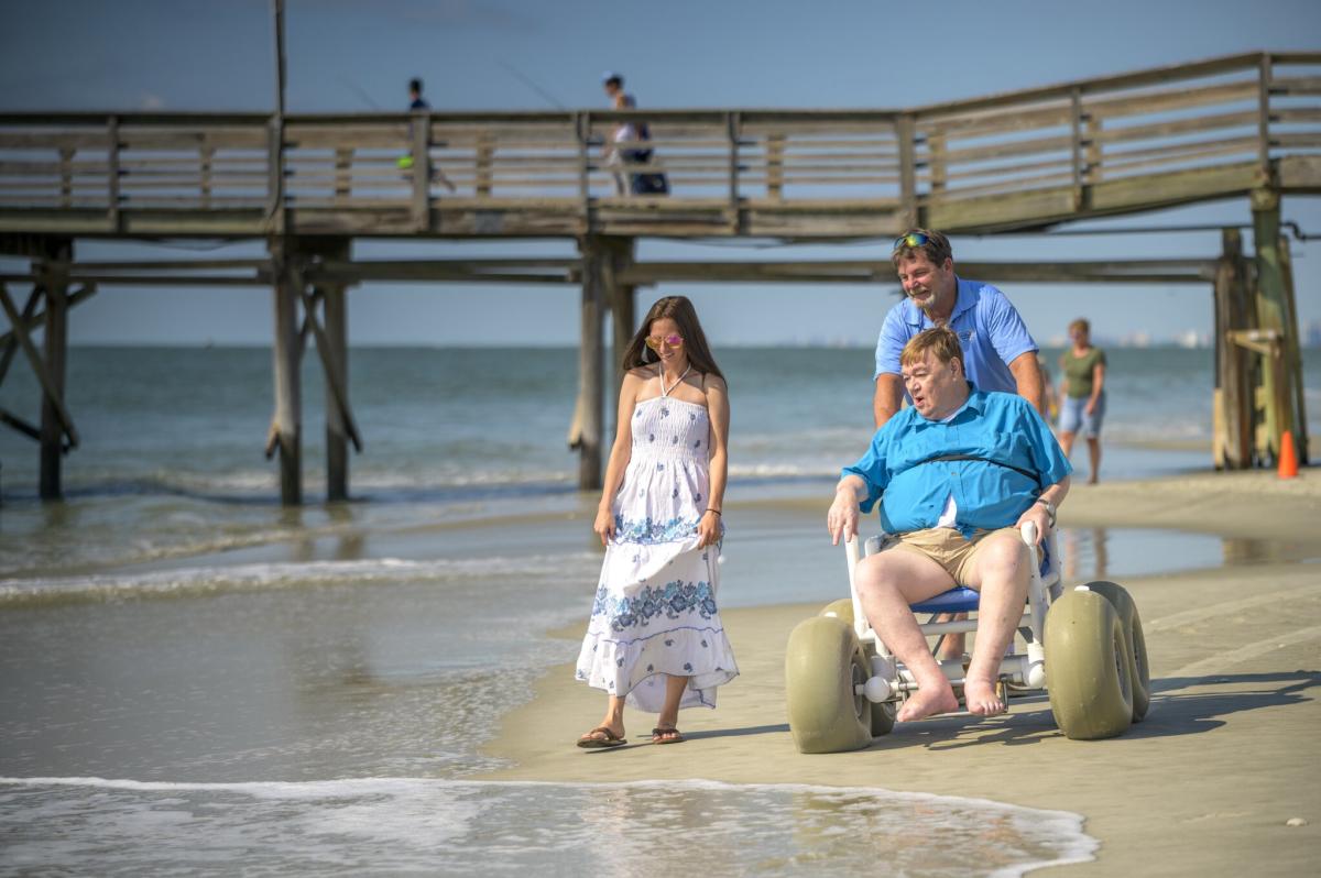A man in a beach wheelchair with large wheels is being pushed along the shore by another man. A woman in a white dress walks beside them. In the background, there is a wooden pier extending into the ocean with a couple of people fishing. The scene is bright and sunny with calm waves on the sandy beach.