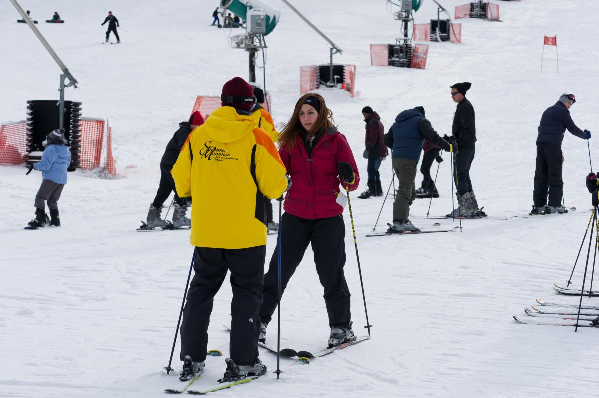 A ski instructor wearing a yellow jacket that reads "Shawnee Mountain" instructs a woman on proper technique