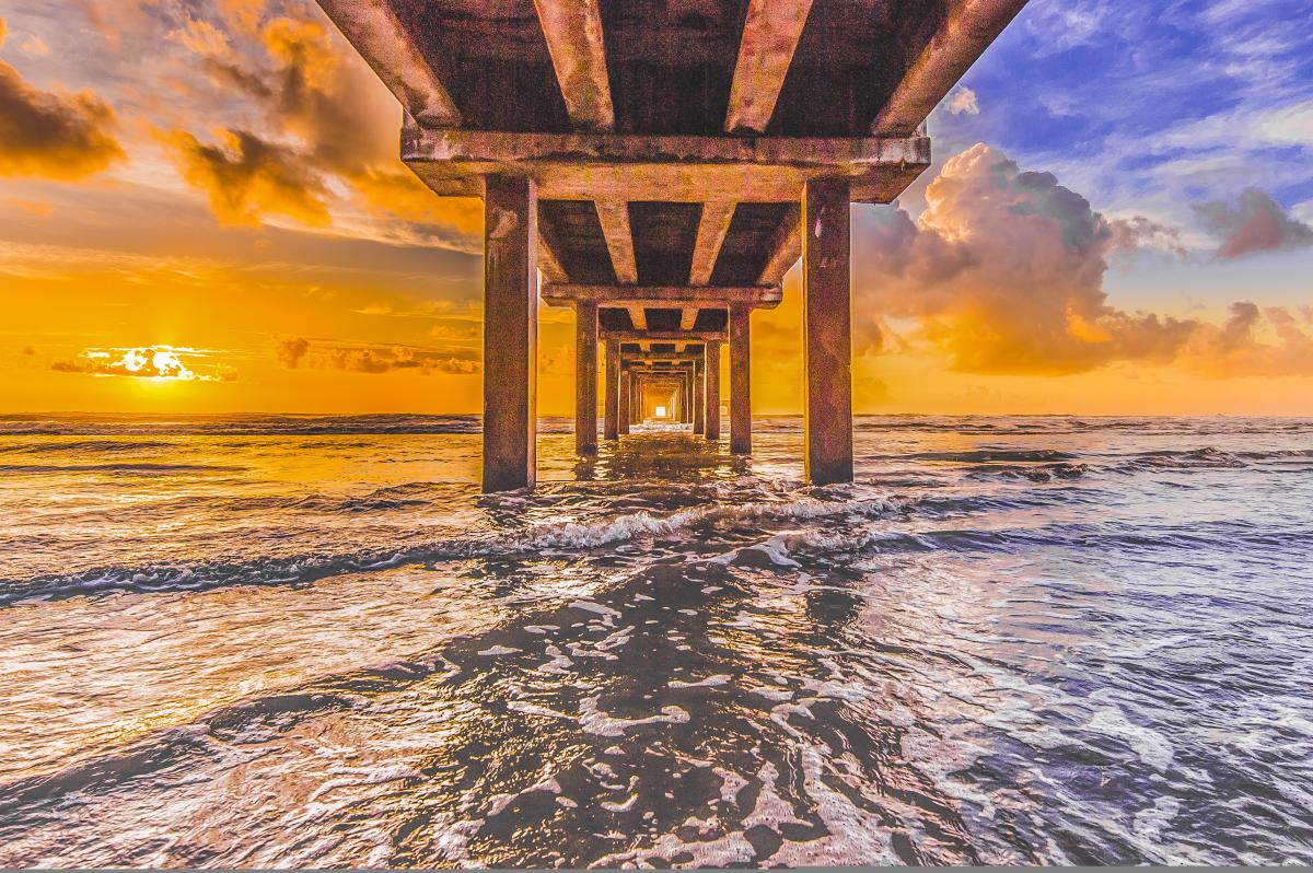 The sun rises just over the water from the perspective of under a pier