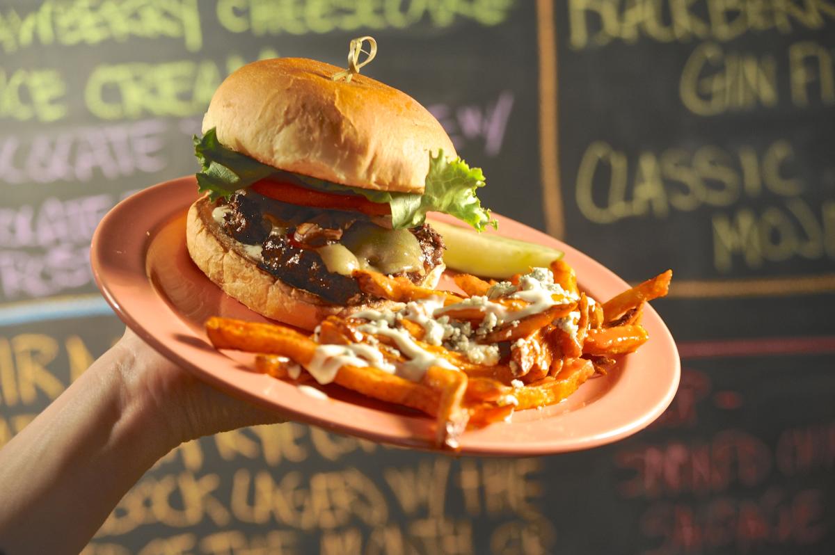A mouthwatering burger with melted cheese, lettuce, tomato, and pickles sits on a bun, accompanied by a side of sweet potato fries topped with crumbled blue cheese and sauce. The plate is held up in front of a chalkboard menu with colorful handwritten text.