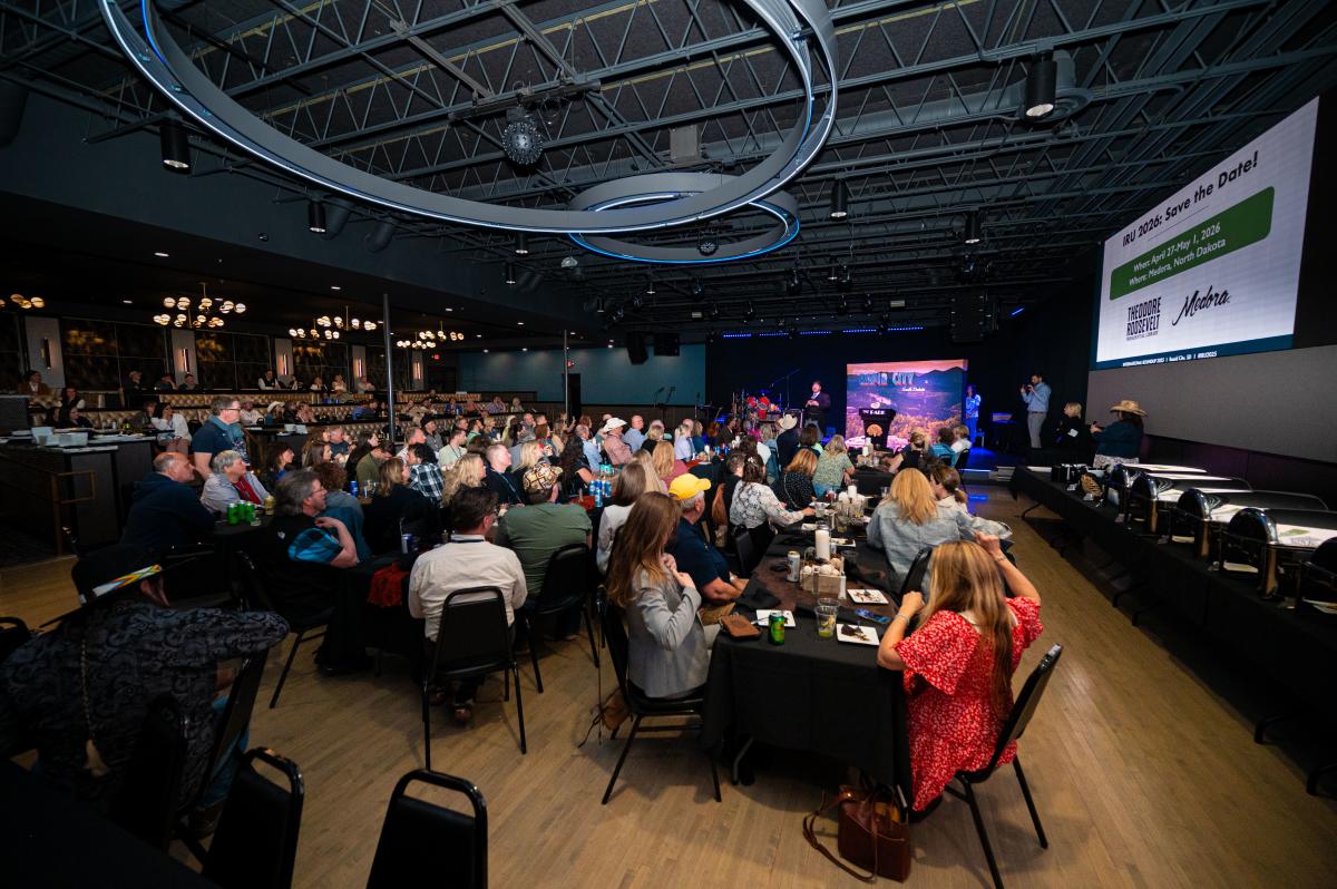 Conference attendees seated at round tables watch a presentation inside The Park, with a buffet, stage, and large event screen visible.