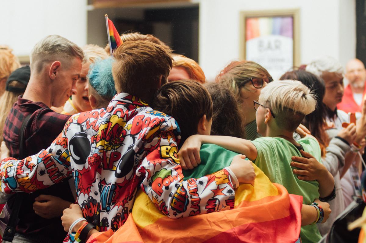 Big group of people embracing, with someone holding a pride rainbow flag