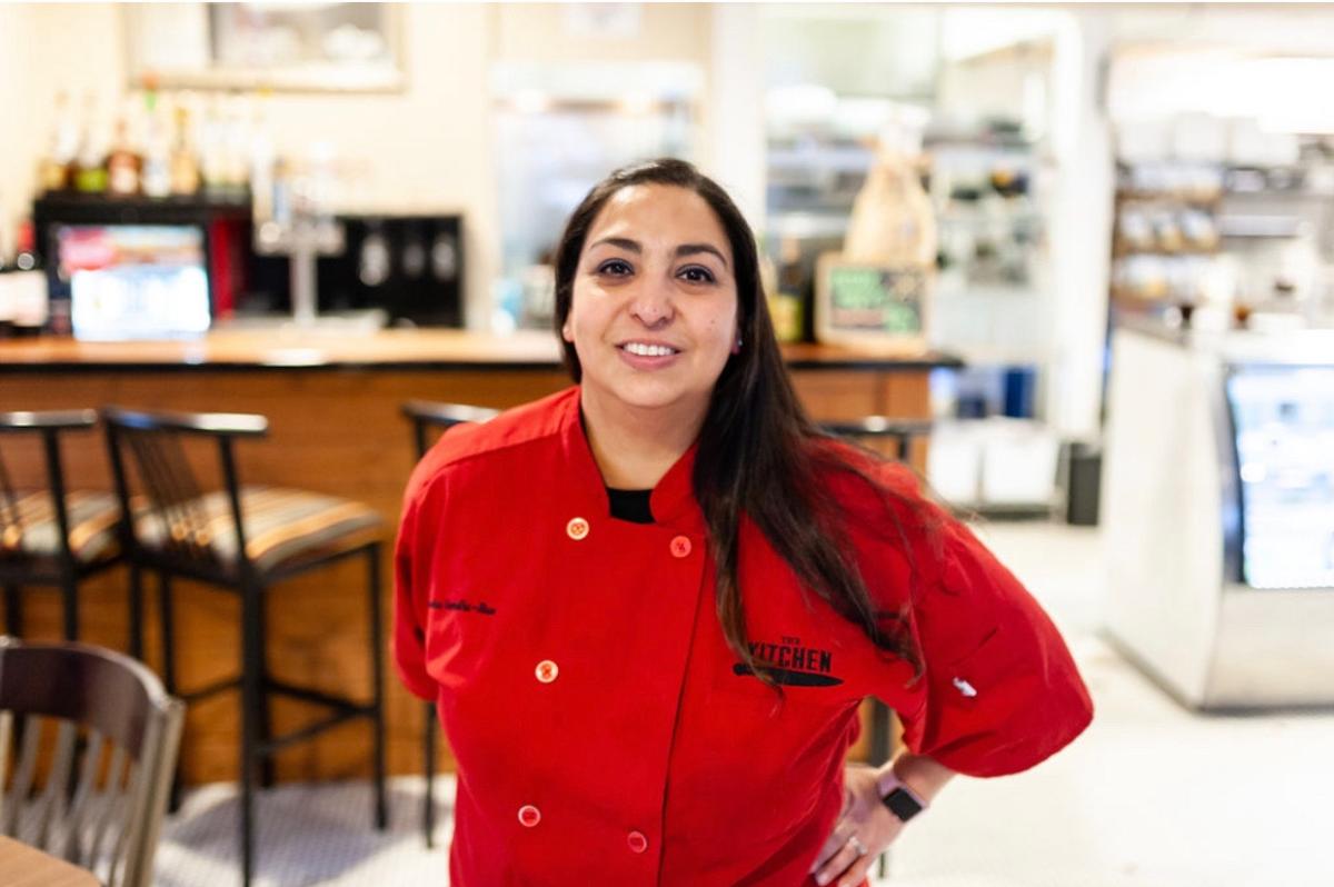 Natasha Gandhi-Rue, chef and co-owner of The Kitchen, poses in her red chef coat in the dining room of her restaurant