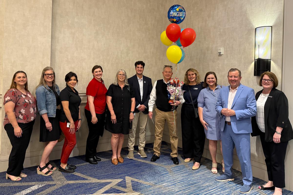 Visit The Woodlands and Jennifer Gohagan award Van Silliman with his 2025 NTTW Hospitality Award at The Woodlands Waterway Marriott. Featured are Amber Parsley, Ashley Fenner, Carla Twidell, Rachael Varner, Julie Quinn, Ryan Greenwood, Van Silliman, Josie Lewis, Elizabeth Eddins, Nick Wolda, and Jennifer Gohagan. Everyone is dressed in business or business casual attire. Van is holding a candy bouquet and primary-colored balloons, one of which reads, "CONGRATS," with shooting stars around it.