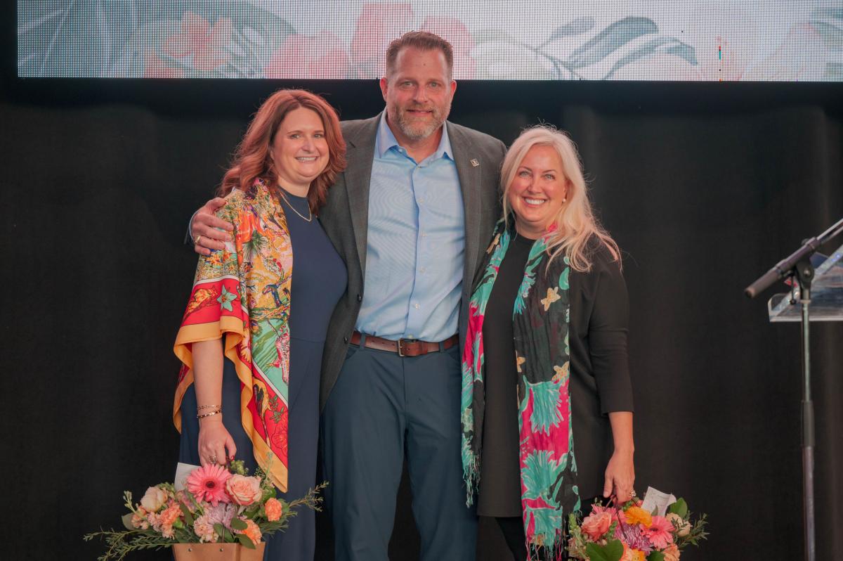 Three people (two women and one man in between them) smile at the camera. They are standing on The Pavilion stage, dressed in business casual wear. The woman on the right has auburn hair and is wearing a navy dress with an orange-red-and-blue shawl depicting a scene of water birds by a pond. The woman on the right has white hair and a young face. She's wearing a long black blouse and black pants with a colorful long scarf hanging loosely from her neck. Both women are carrying small metal buckets filled with bouquets of pink and orange flowers. The man in the middle has his arms around both women. He's wearing a gray blazer with a light blue shirt and blue-gray slacks.