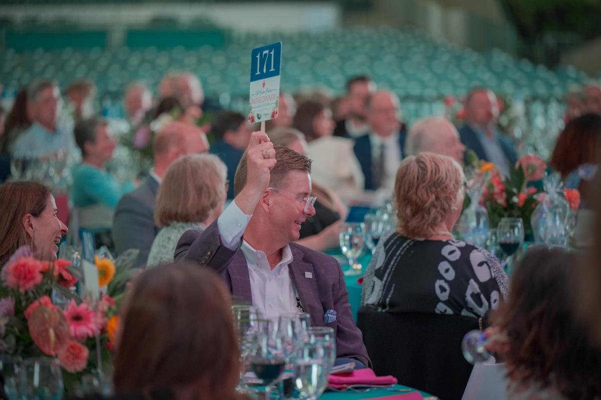 The camera focuses on one man among all the guests of The Pavilion Partners Annual Wine Dinner and Auction. He is looking off to the right of the frame, presumably toward The Pavilion stage, raising an auction bidding paddle. He is wearing clear-framed glasses, a dark gray blazer, and white button-down shirt with an A&M logo under one lapel. The paddle says "171" in large font at the top, and below it, "The Pavilion Partners WINE DINNER AND AUCTION." There is smaller text below it that can't be read from the camera's distance.