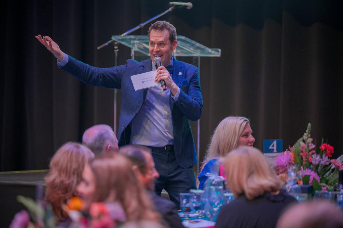 A man in a royal blue blazer, light blue button-down shirt, and navy slacks speaks into a microphone while raising an arm to address the dinner guests. In the same hand as the microphone, he is holding a stack of index cards that appear to read, "PatTully.com" and "@PatrickJTully" beside an Instagram logo. Six guests are visible but out of focus in front of him, all seated at a round, blue-clothed table with a pink-and-orange floral centerpiece.