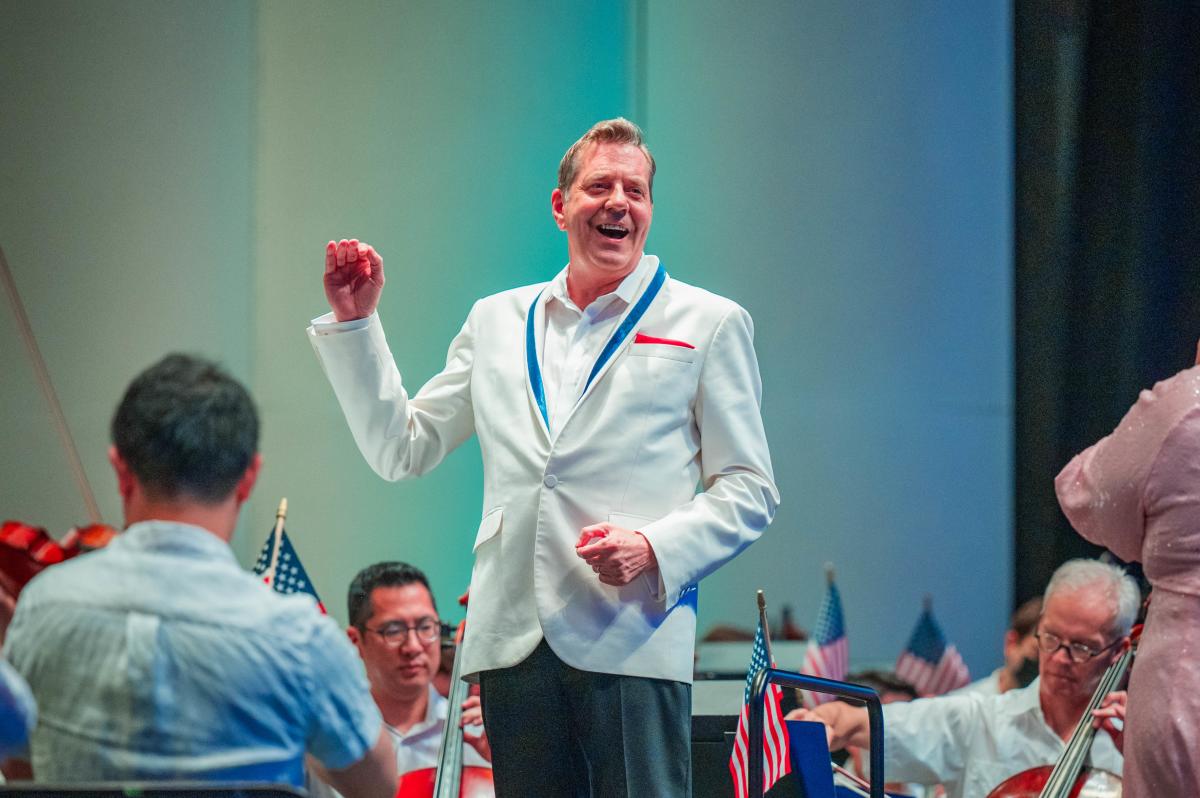 A conductor in a white blazer with red and blue trim instructs his orchestra at The Cynthia Woods Mitchell Pavilion. The walls are lit with blue lights, and American flags are propped up around the stage.