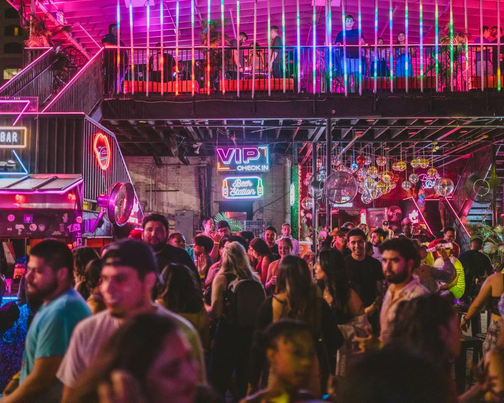 Crowd of people standing on an outdoor patio covered in hot pink, yellow, blue and green neon lights.