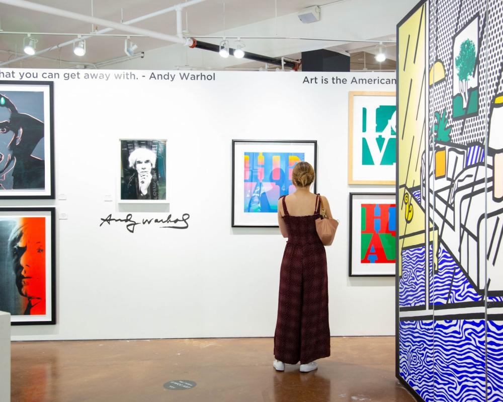 A woman standing in the middle of an art gallery looking at a piece of art mounted on a white gallery wall.