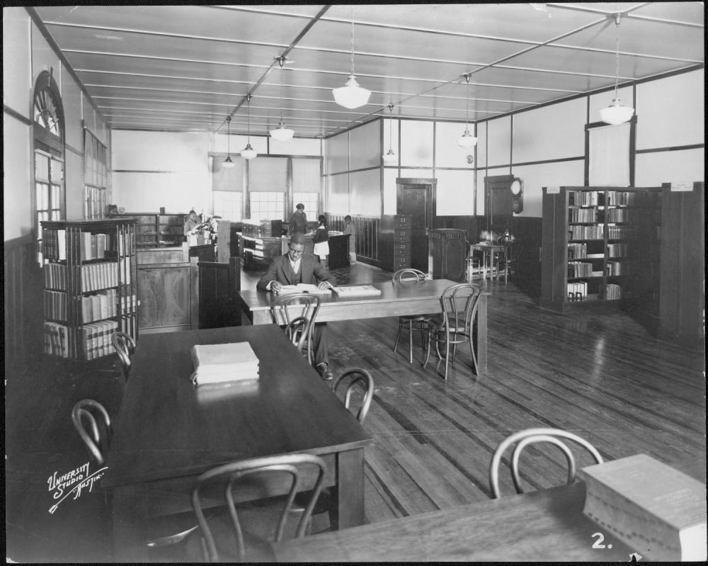 A Black man wearing glasses is looking at a book whlie sitting at a table inside the Carver Branch Library. It is a wide view of the room and there are a couple of children checking out books in the background