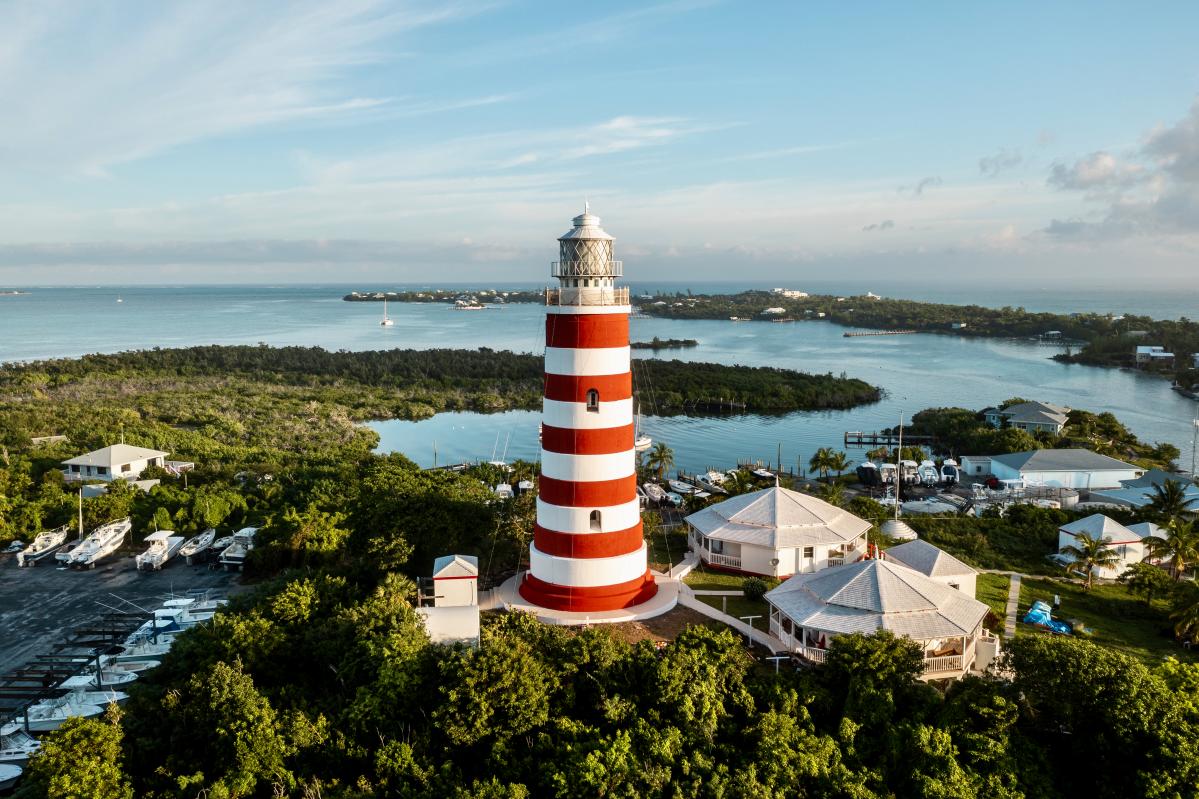 An aerial view of the iconic red and white striped lighthouse on Hog Island in the Bahamas.  The lighthouse stands tall against a backdrop of turquoise water and lush greenery. Boats are docked nearby, and a small village is visible in the distance.