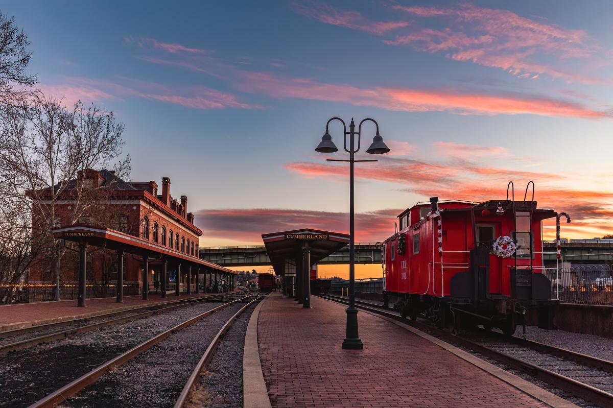 A red train caboose with a holiday wreath sits parked on train tracks in front of a historic train station at sunset.