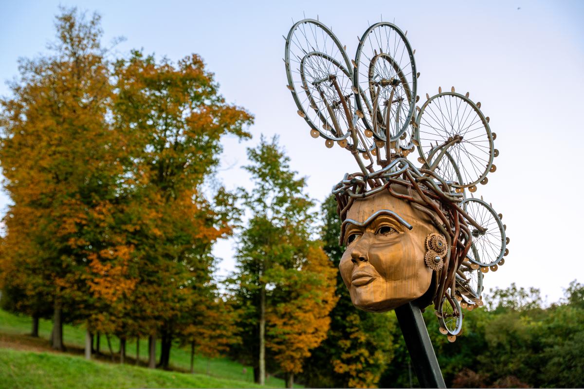 The art sculpture featuring a golden face with bike wheels as hair is the focus with tree changing to fall colors in the background.