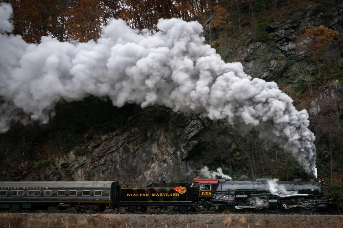 The Western Maryland Scenic Railroad steam engine billows smoke from the engines and pulls along a passenger car near a mountain wall.